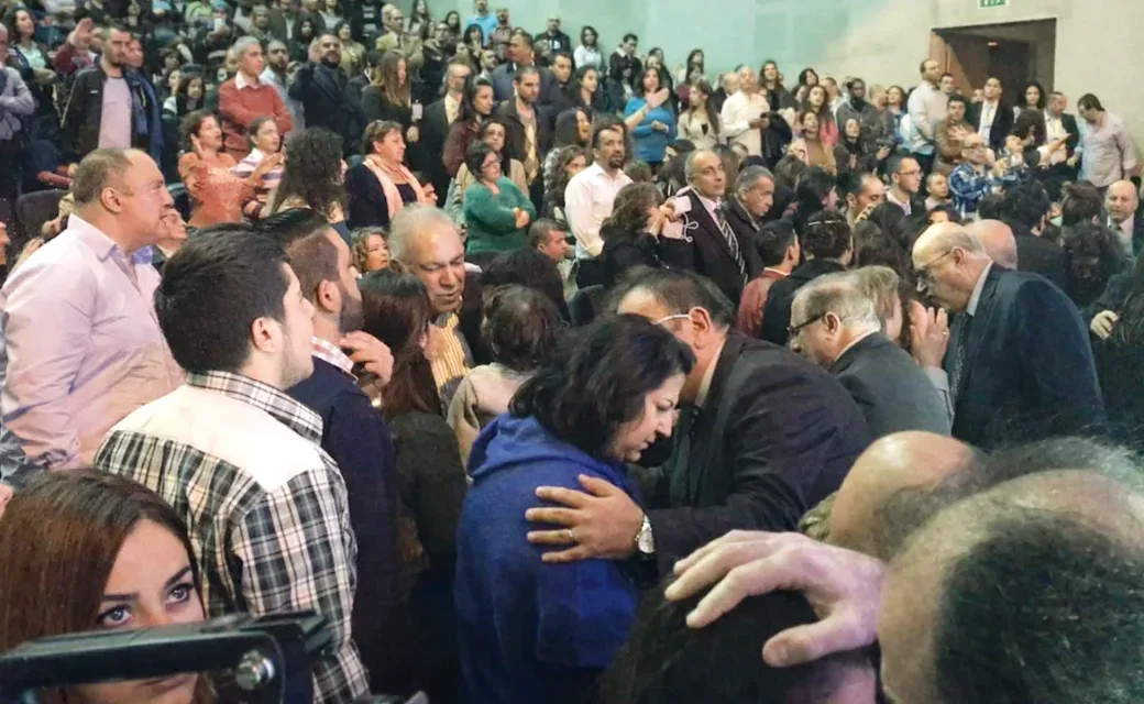Crowded indoor event in Beirut with people standing in aisles while others pray and comfort one another.