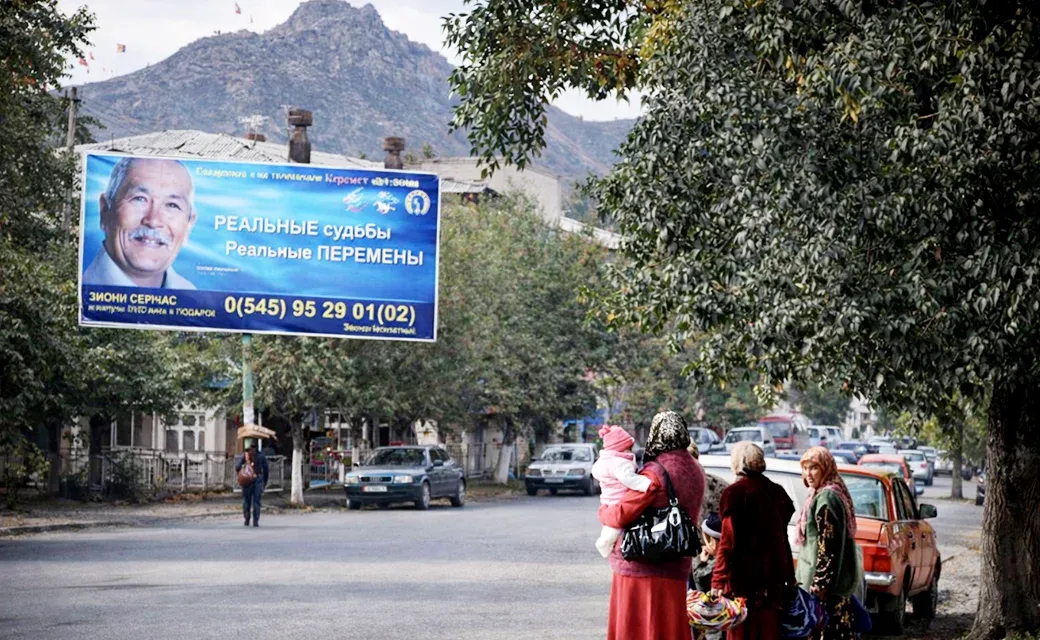 People walk along a busy street in the Fergana Valley while a large billboard with a man’s face and a phone number stands above the road.