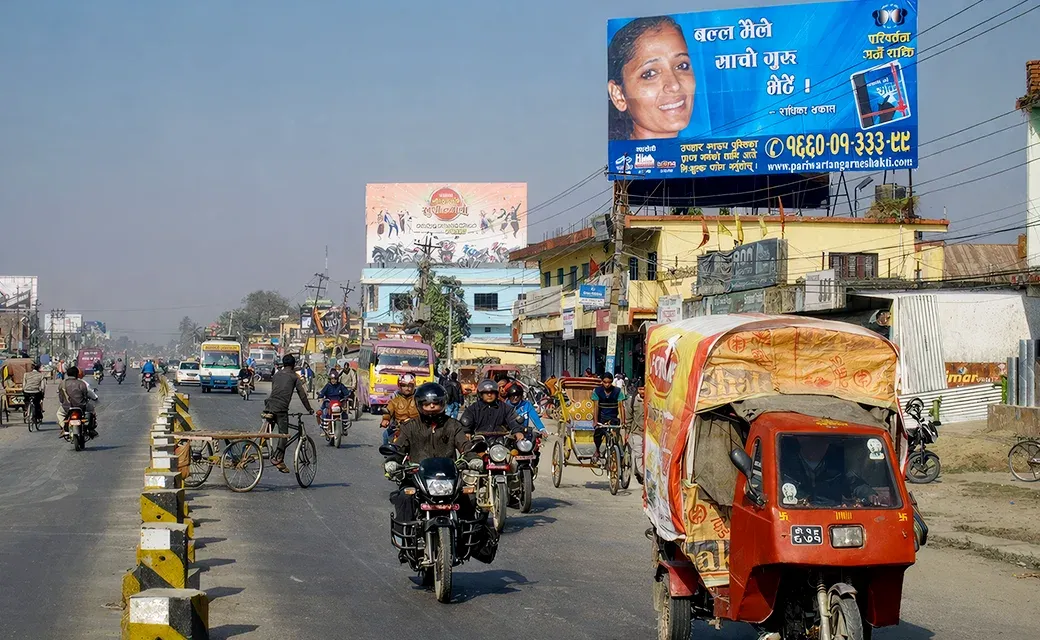 A busy road with motorbikes and cars passing beneath a large roadside billboard featuring a man’s face and campaign text.