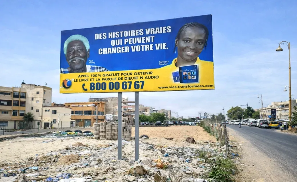 Street scene in Dakar, Senegal, with cars and motorbikes passing an outdoor roadside billboard.