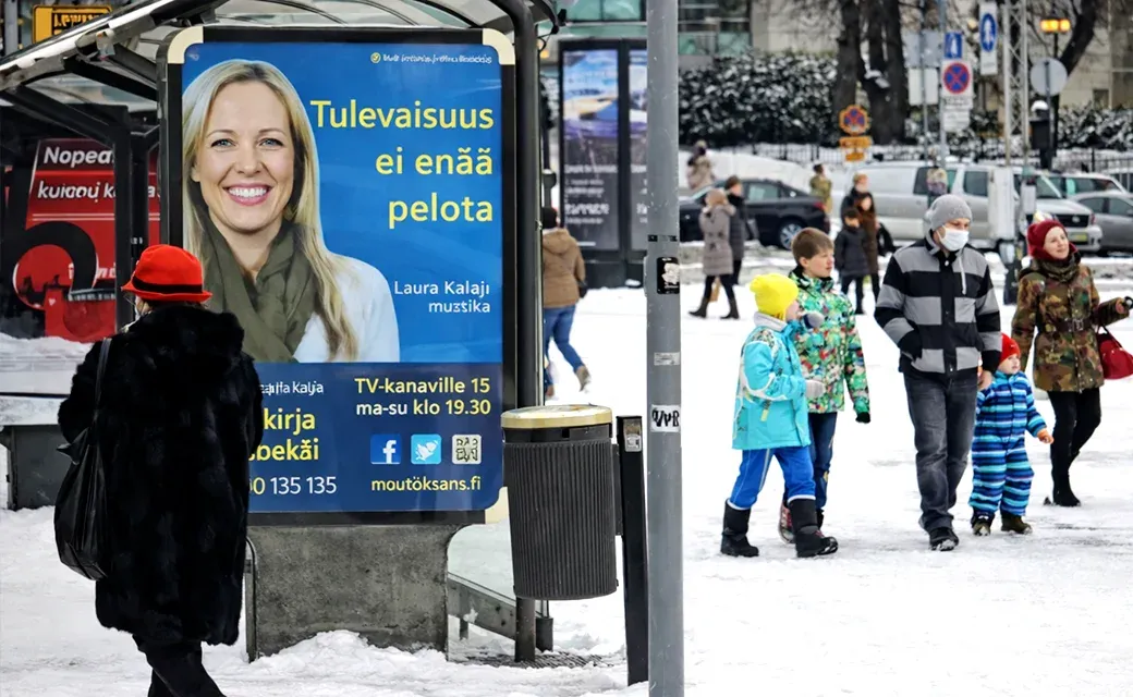 People walking in a snowy city square beside a bus shelter showing a Finnish-language campaign poster.