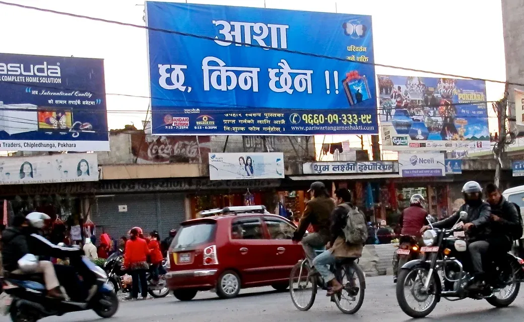 A crowded marketplace in Kathmandu with a large campaign advertisement visible in the background promoting a free book about Jesus.