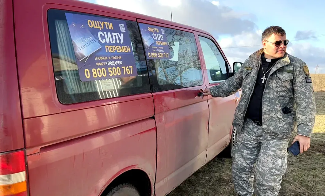 A man in military-style clothing stands beside a red van with two campaign posters displayed in the side windows.