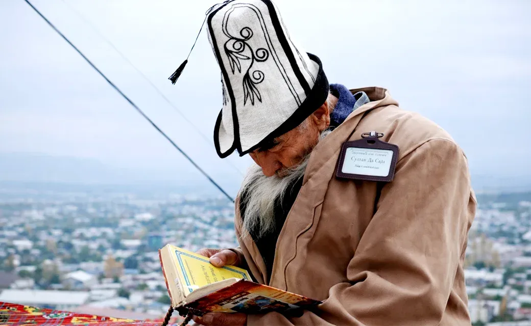 Elderly man in a traditional Kyrgyz hat reading a book outdoors with the city of Osh in the background.