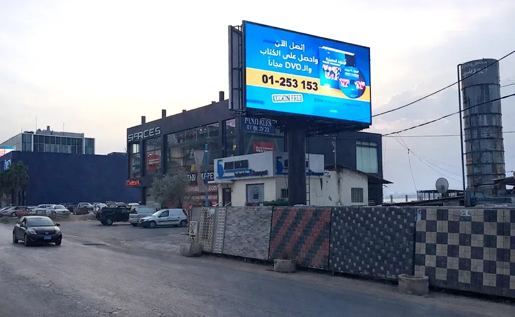 A busy road with motorbikes and cars passing beneath a large roadside billboard featuring a man’s face and campaign text.