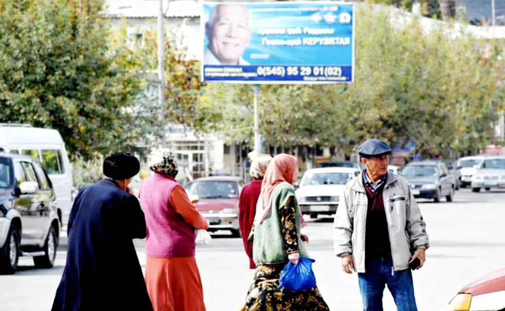 People crossing a street in Kyzyl-Kiya, Kyrgyzstan with a blue campaign billboard in the background.