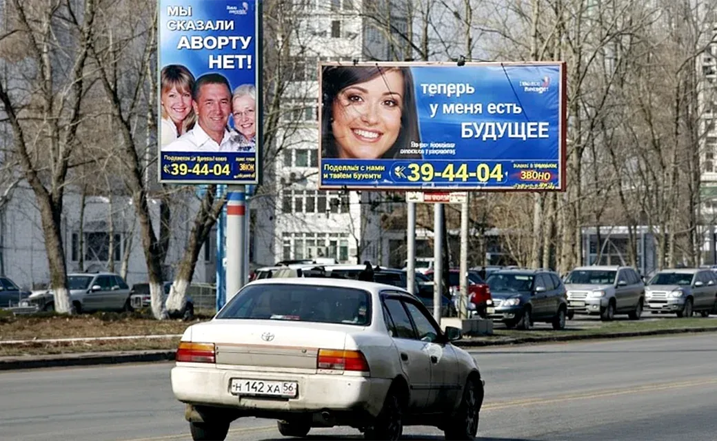 People waiting at a street stop near a Russian-language poster