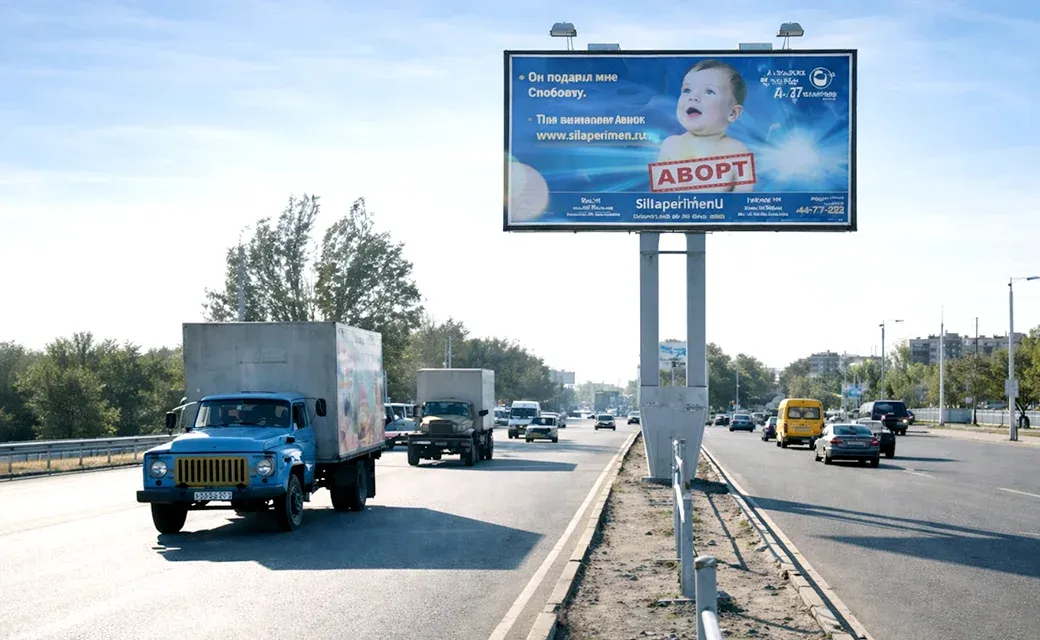 Roadside billboard with a baby photo and Kyrgyz/Russian text, including a call-in phone number, above a busy highway.