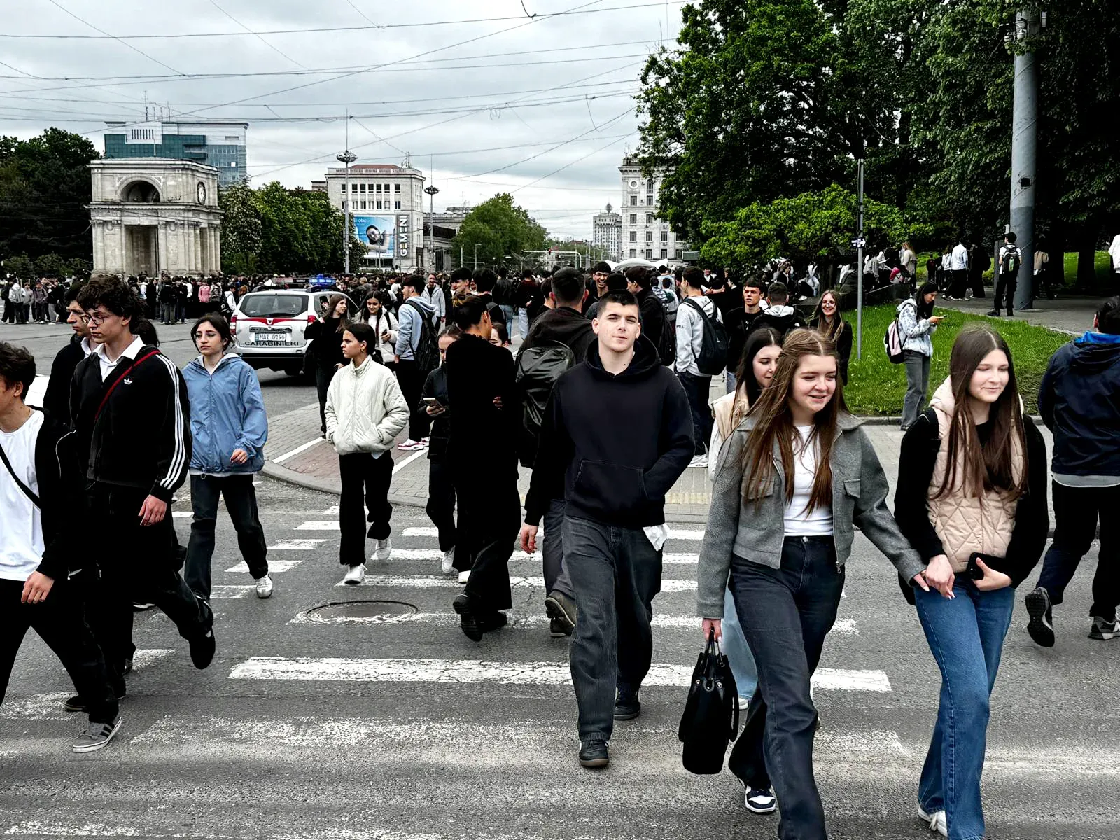 Crowds of people cross a city street in Chisinau, Moldova, with historic buildings and trees in the background.