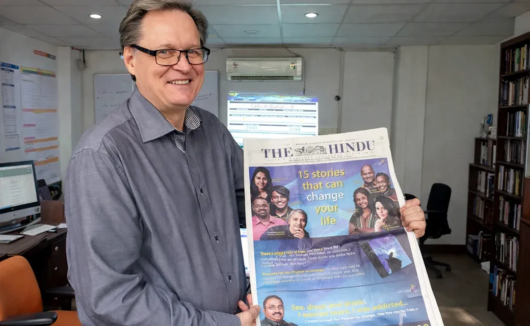 Hannu Haukka holding a copy of The Hindu newspaper with a large campaign advertisement on the front page; a call-center screen is visible behind him.