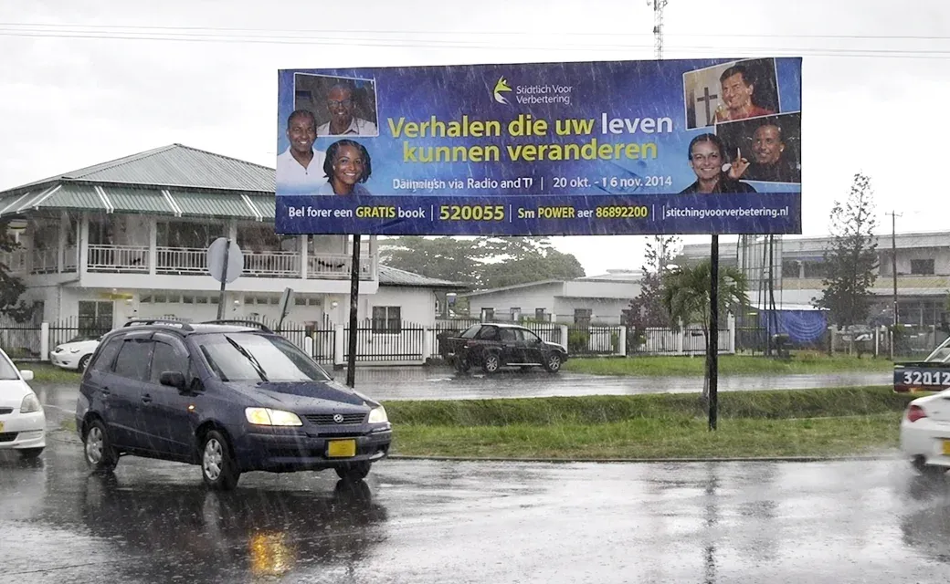 Roadside billboard in Paramaribo promoting Power to Change Suriname, photographed during rainfall with cars passing.