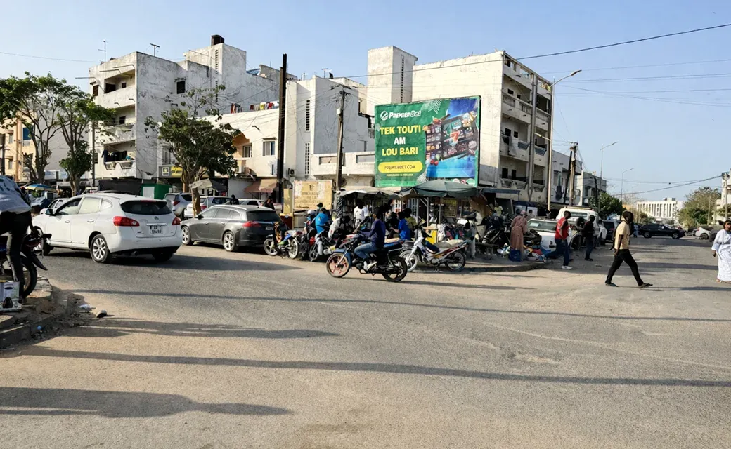 Street scene in Dakar, Senegal, with cars and motorbikes passing an outdoor roadside billboard.