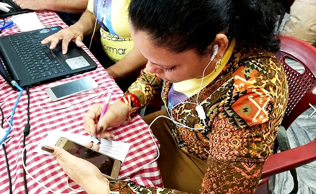 Woman wearing earphones writes on a form at a table beside a laptop and phones.