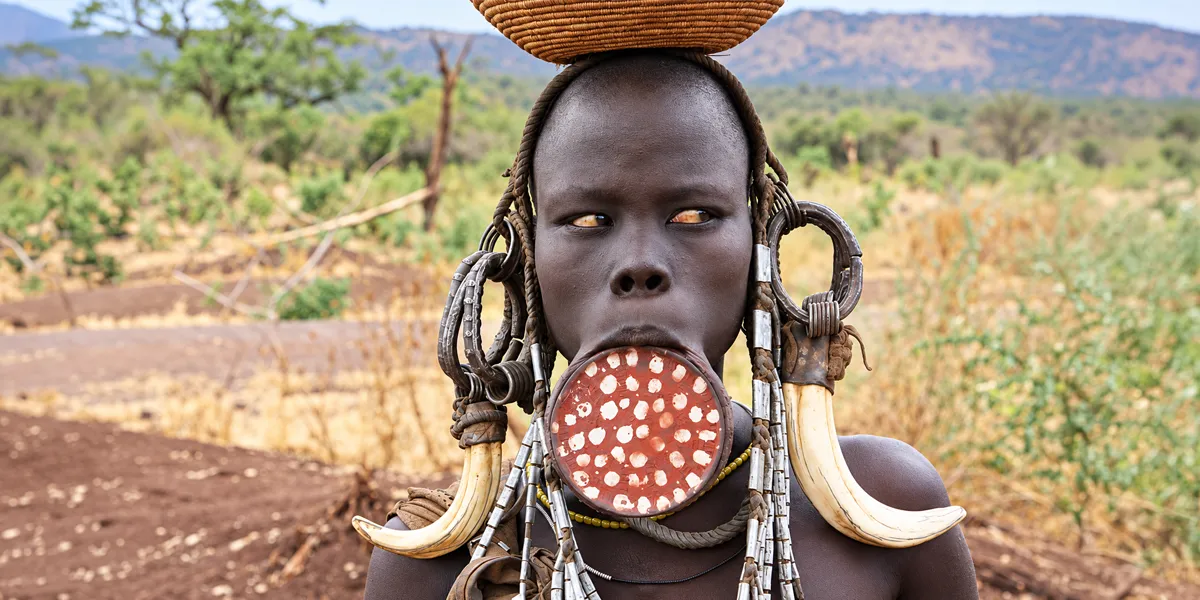 Mursi woman in southern Ethiopia wearing a large lip plate and horn ear ornaments, with a woven basket on her head