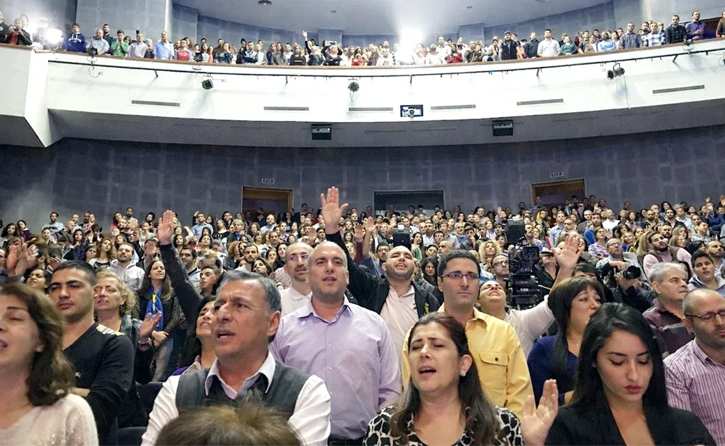 Large indoor auditorium in Beirut filled with people; many stand and raise their hands during a worship moment.