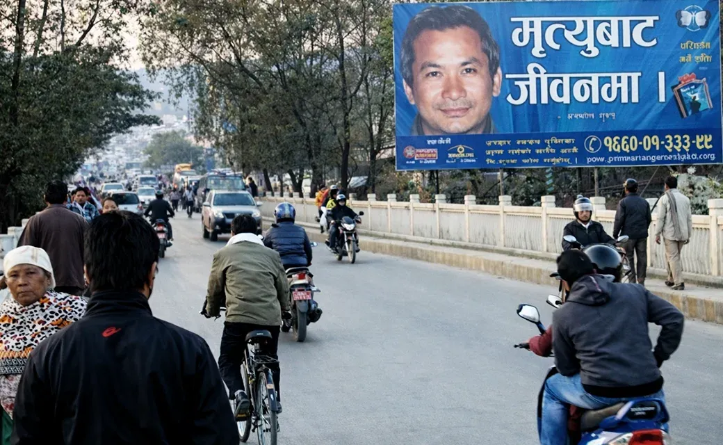 A busy road with motorbikes and cars passing beneath a large roadside billboard featuring a man’s face and campaign text.