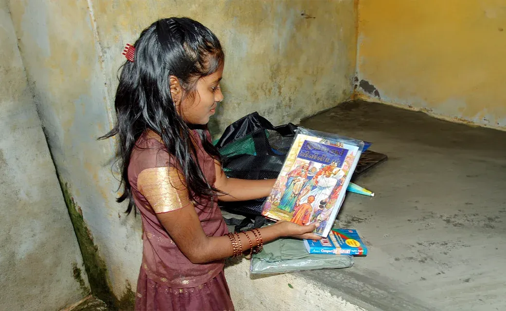 A young girl holds up a children’s picture Bible beside a backpack and school supplies indoors.