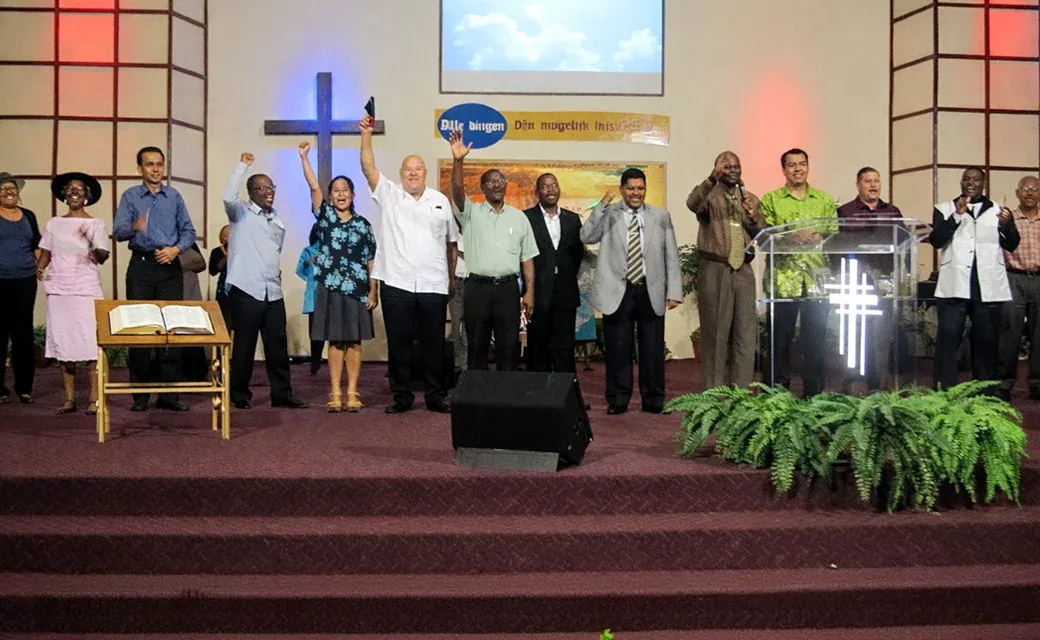 Group of church leaders standing on a stage in a church sanctuary, raising hands near a podium and cross.