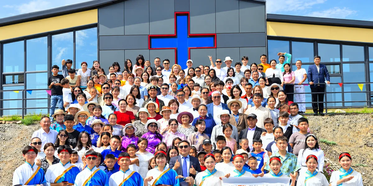 A large group of adults and children gathers outside a church building in Mongolia, posing beneath a large blue cross outlined in red.