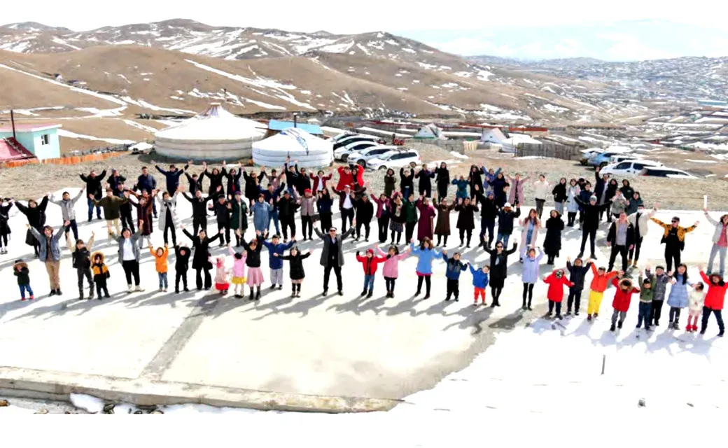 A large group of adults and children waving together outdoors in Mongolia, with traditional gers, buildings, and snow-covered mountains in the background.