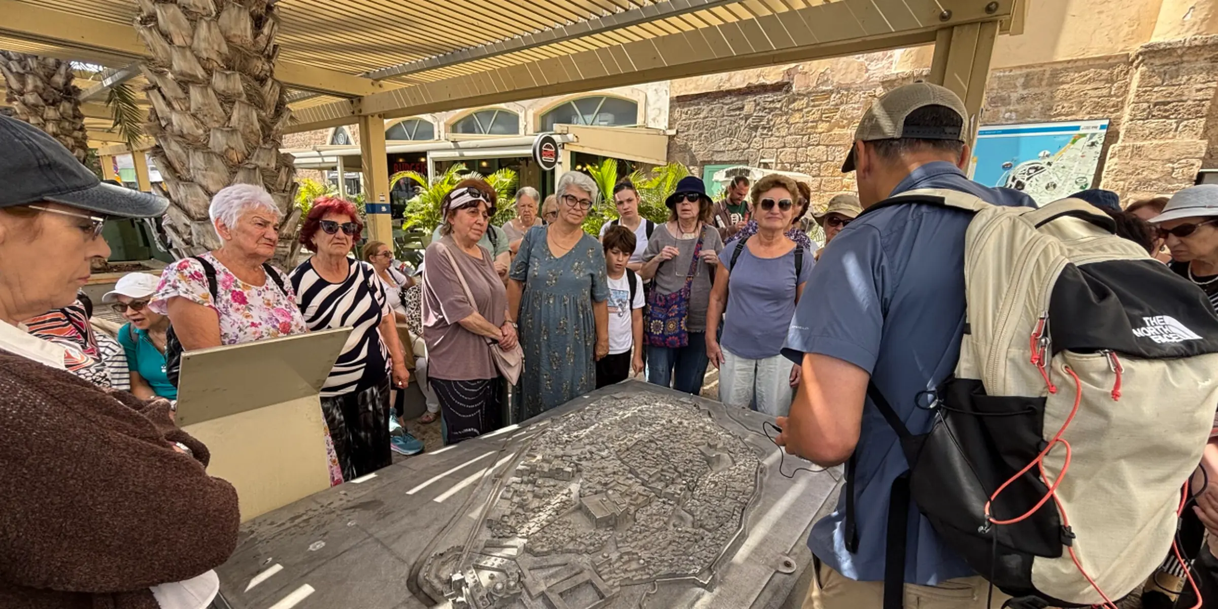 A group of people gathers around a large raised relief map beneath a shaded outdoor structure, while a man wearing a backpack stands facing the group.