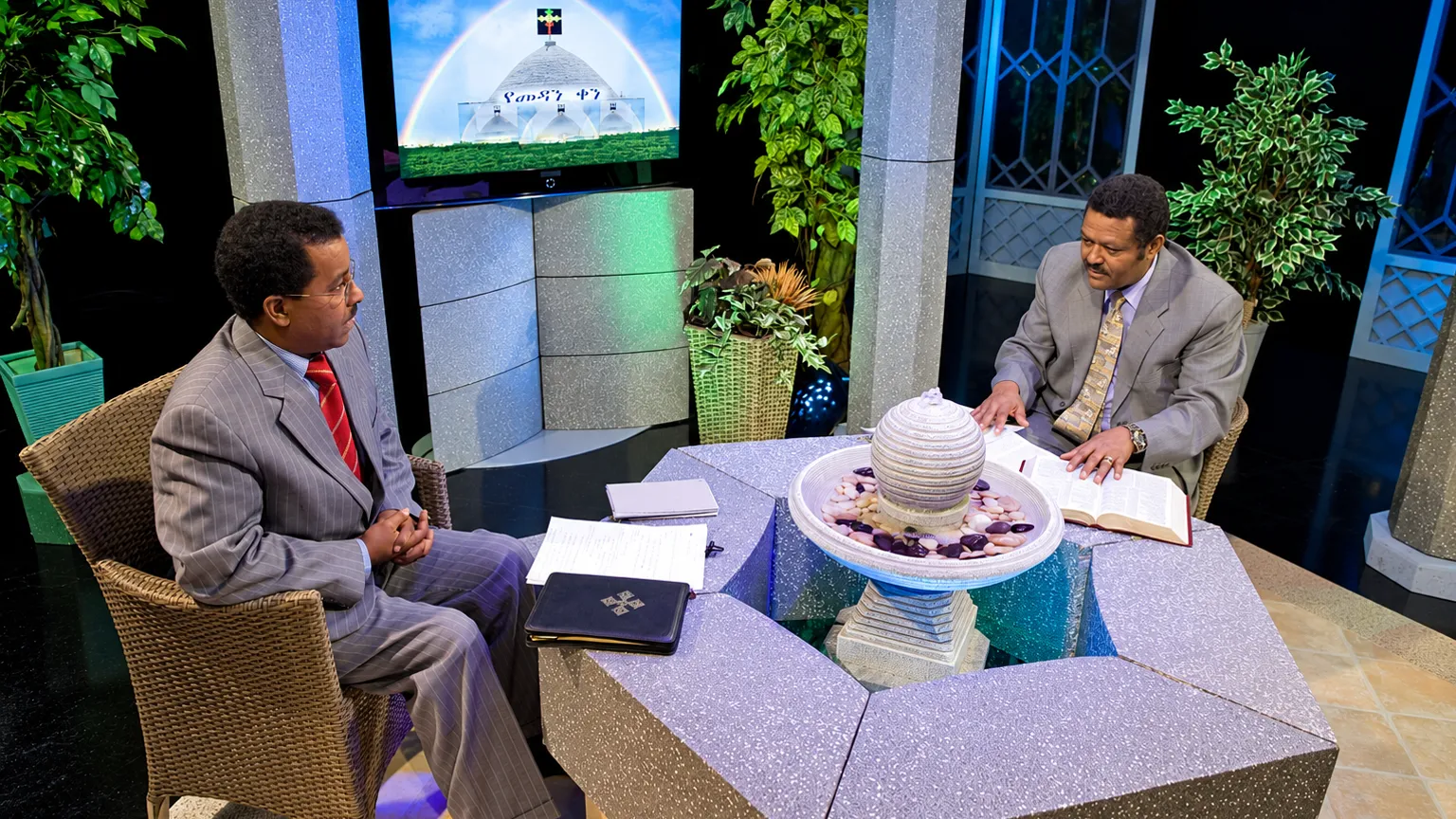 Two men discussing Bible teachings in a Christian television studio, with an open Bible on the table.