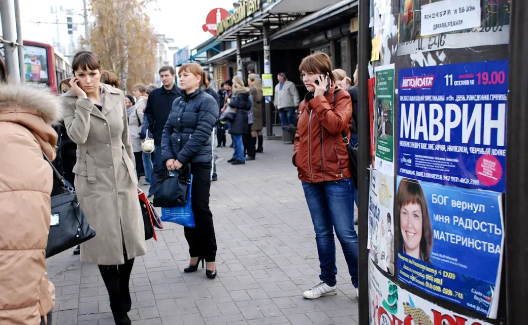 People waiting at a street stop near a Russian-language poster