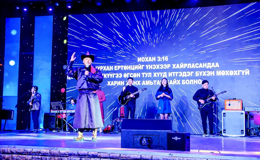 A Mongolian worship band performs on a brightly lit stage, with singers and musicians leading worship during a gospel event.
